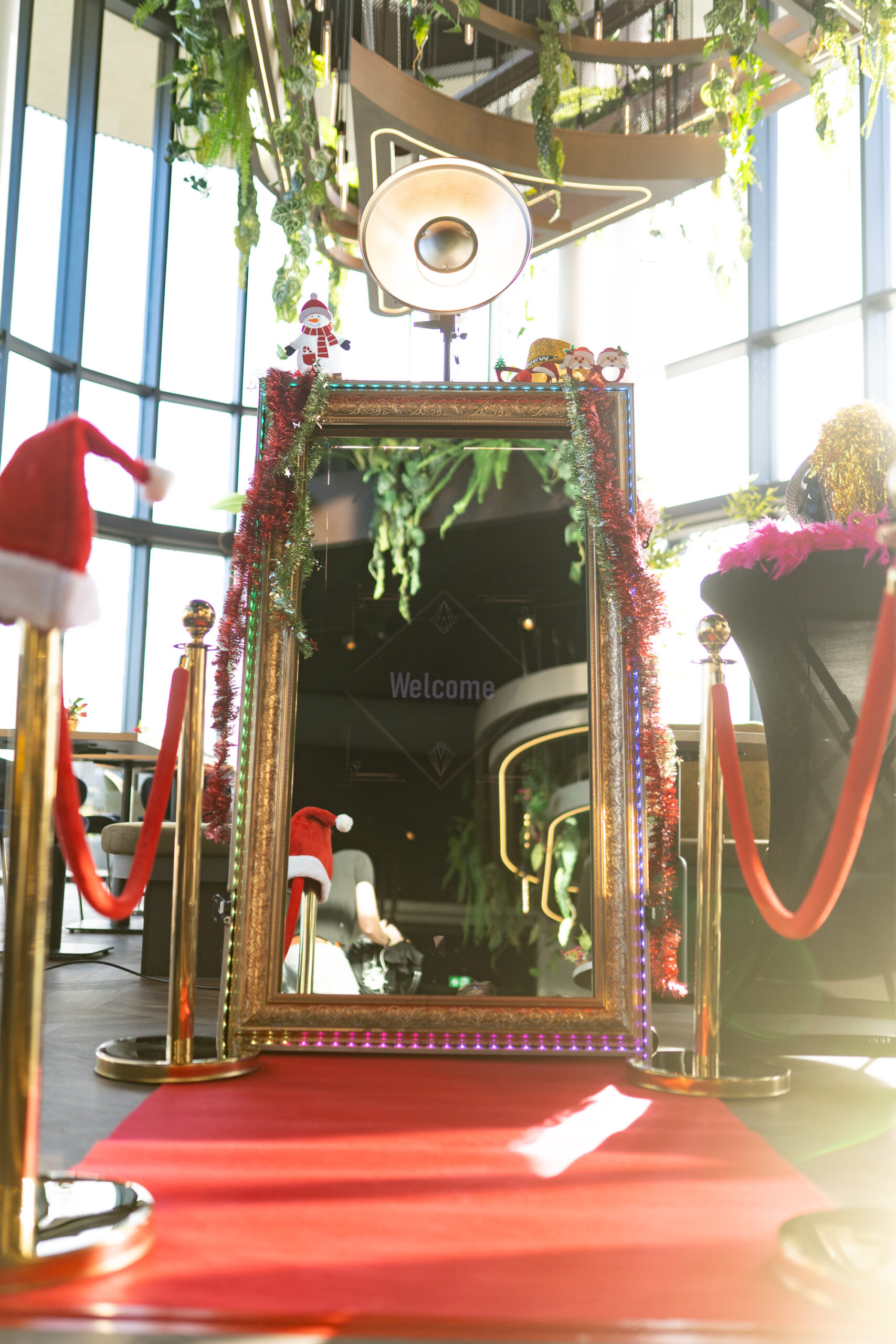 Spiegel photobooth in volledige opstelling: gouden sierlijst met verlichte LED-rand, uitgerold op een rode loper tussen gouden koordpaaltjes, feestelijk versierd met kerstslingers.